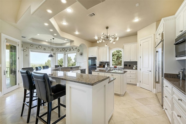 kitchen featuring an inviting chandelier, pendant lighting, a raised ceiling, white cabinetry, and a kitchen island with sink