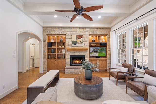 living room featuring built in shelves, beamed ceiling, a fireplace, ceiling fan, and light hardwood / wood-style flooring