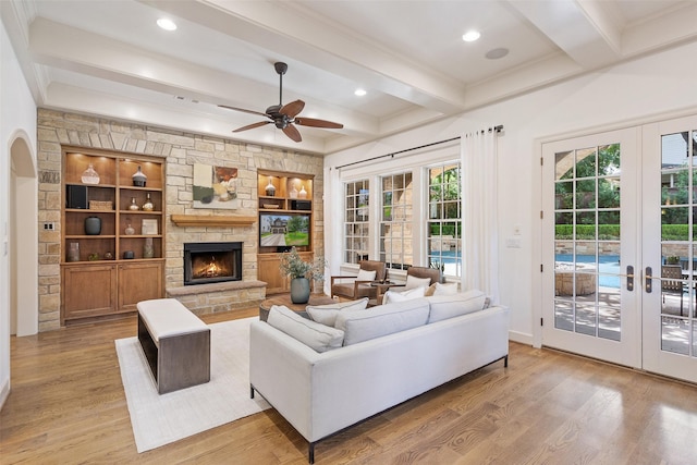 living room featuring ceiling fan, light hardwood / wood-style flooring, built in features, a stone fireplace, and french doors