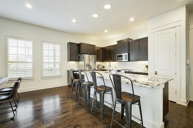 kitchen with light stone counters, a breakfast bar area, a center island with sink, stainless steel appliances, and dark wood-type flooring