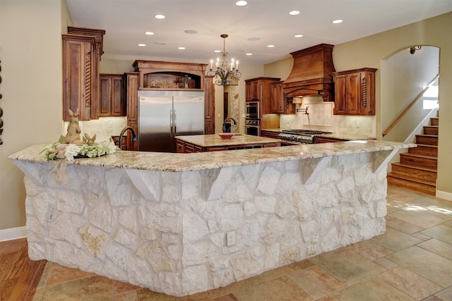 kitchen featuring a kitchen island with sink, hanging light fixtures, backsplash, stainless steel appliances, and custom exhaust hood