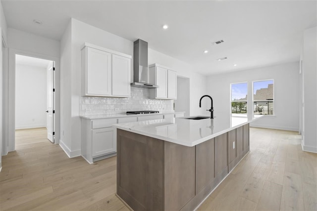 kitchen with light wood-type flooring, white cabinetry, sink, wall chimney exhaust hood, and a kitchen island with sink