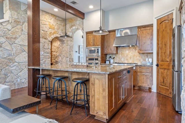 kitchen with hanging light fixtures, dark hardwood / wood-style flooring, stainless steel appliances, light stone countertops, and wall chimney exhaust hood