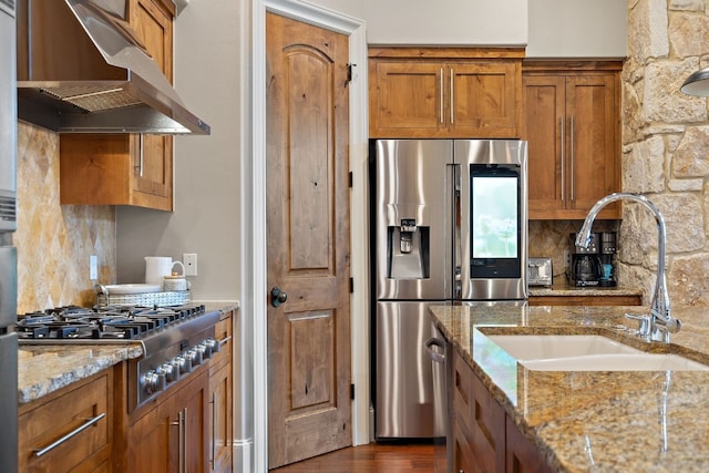 kitchen with backsplash, sink, stainless steel appliances, light stone counters, and wall chimney range hood