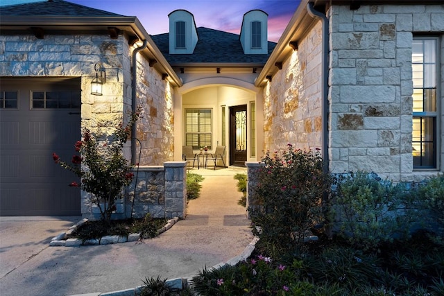 138 Loysoya St, Bastrop, TX 78602 - exterior entry at dusk featuring stone siding, an attached garage, a shingled roof, and a porch