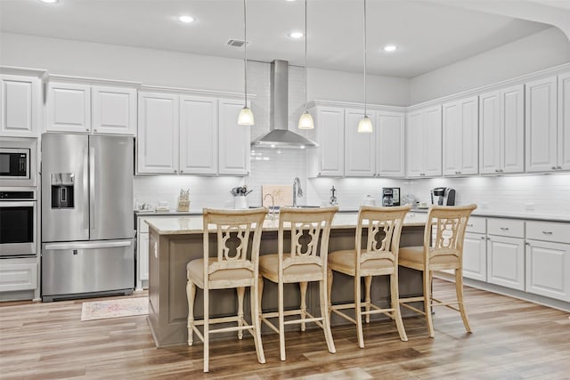 kitchen featuring white cabinets, an island with sink, wall chimney range hood, pendant lighting, and appliances with stainless steel finishes