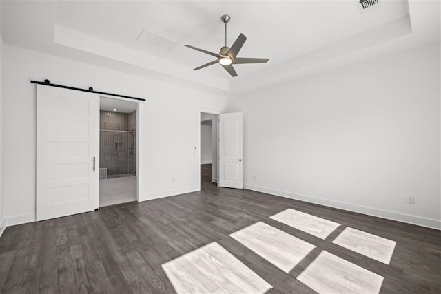 unfurnished bedroom with ceiling fan, a barn door, connected bathroom, a tray ceiling, and dark hardwood / wood-style flooring