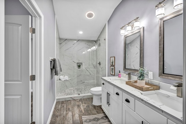 bathroom with dual bowl vanity, toilet, a tile shower, and wood-type flooring