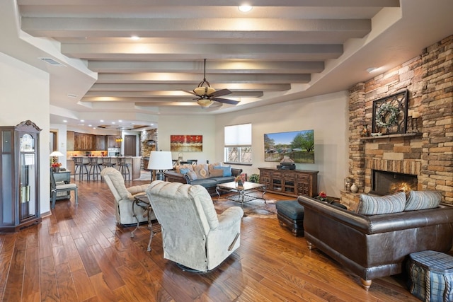 living room with beam ceiling, a stone fireplace, dark hardwood / wood-style floors, and ceiling fan