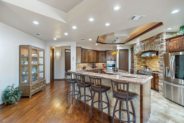 kitchen with stainless steel appliances, light stone countertops, backsplash, a tray ceiling, and a kitchen island with sink