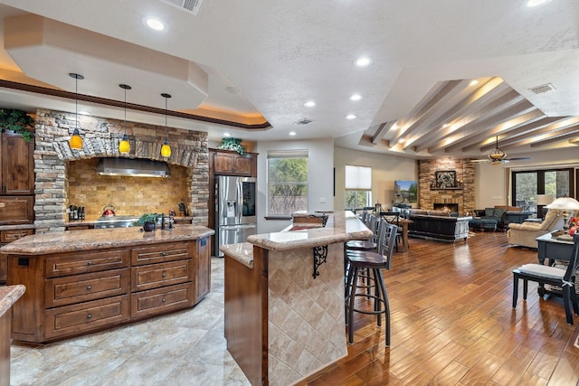 kitchen featuring a kitchen island, ceiling fan, stainless steel fridge with ice dispenser, a stone fireplace, and a tray ceiling