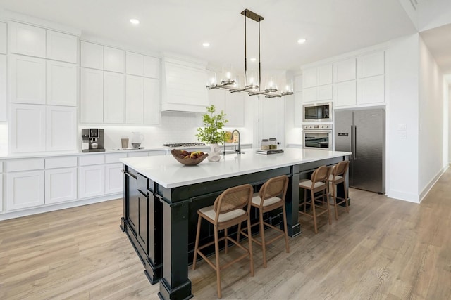 kitchen featuring appliances with stainless steel finishes, an island with sink, hanging light fixtures, white cabinetry, and light hardwood / wood-style floors