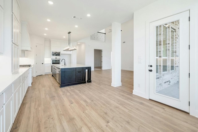 kitchen with appliances with stainless steel finishes, light wood-type flooring, an island with sink, hanging light fixtures, and white cabinetry