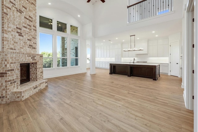 unfurnished living room featuring a towering ceiling, sink, a brick fireplace, ceiling fan, and light hardwood / wood-style flooring