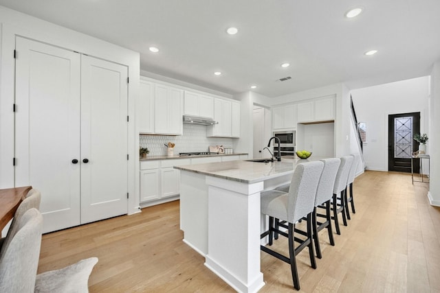 kitchen with light stone counters, stainless steel appliances, light hardwood / wood-style floors, white cabinetry, and a center island with sink