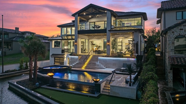 back house at dusk with a balcony, a patio area, and a swimming pool with hot tub