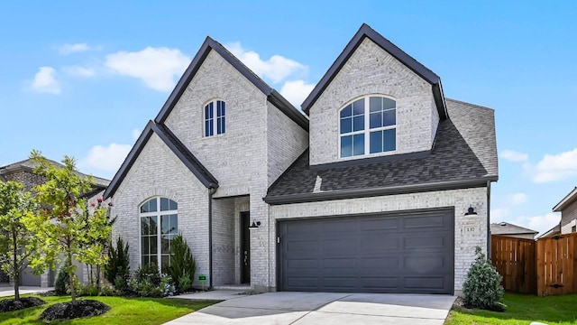 french country inspired facade featuring driveway, brick siding, a shingled roof, and fence