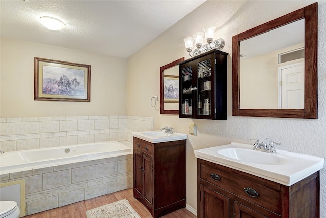 bathroom featuring tiled tub, a textured ceiling, toilet, hardwood / wood-style flooring, and dual bowl vanity