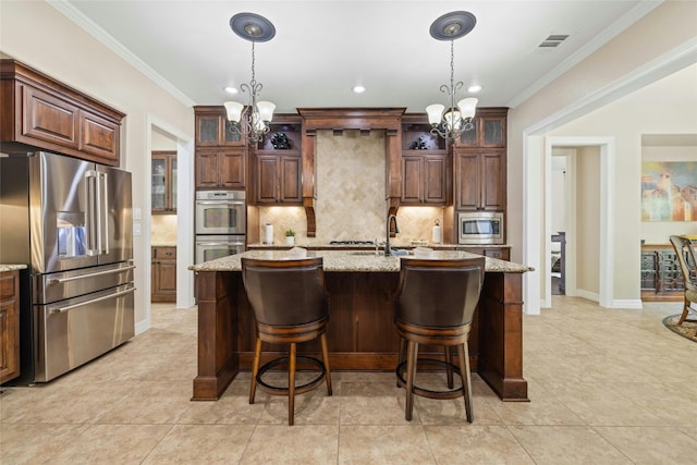 kitchen with appliances with stainless steel finishes, an island with sink, a chandelier, light stone counters, and tasteful backsplash