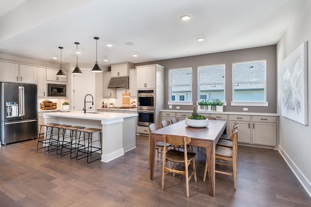 kitchen with appliances with stainless steel finishes, a center island with sink, hanging light fixtures, and dark wood-type flooring