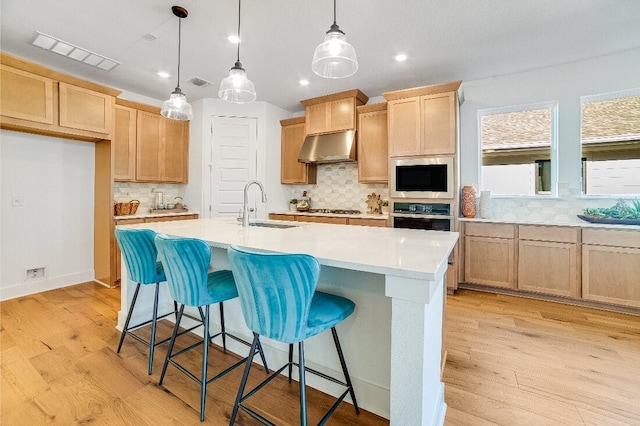 kitchen with light brown cabinetry, built in microwave, sink, hanging light fixtures, and a kitchen island with sink