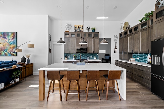 kitchen with decorative backsplash, a kitchen island with sink, black fridge, hanging light fixtures, and light stone countertops