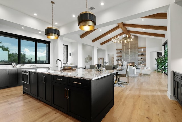 kitchen featuring light stone counters, a kitchen island with sink, vaulted ceiling with beams, light hardwood / wood-style floors, and pendant lighting