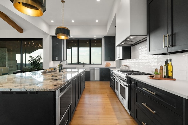 kitchen featuring light stone counters, light hardwood / wood-style floors, stainless steel appliances, custom range hood, and a kitchen island with sink