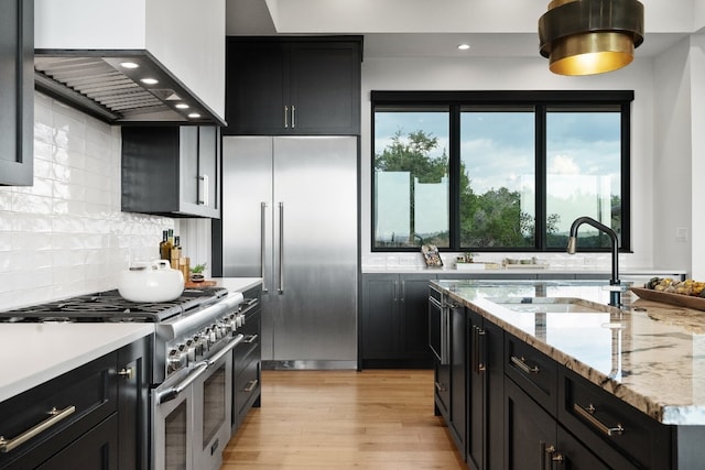 kitchen with custom exhaust hood, backsplash, light stone counters, high quality appliances, and light hardwood / wood-style floors