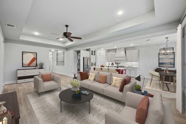 living room with ceiling fan with notable chandelier, sink, a raised ceiling, and light wood-type flooring