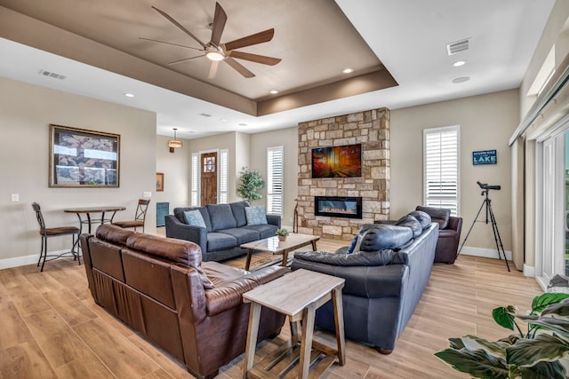 living room featuring a stone fireplace, light hardwood / wood-style flooring, ceiling fan, and a raised ceiling