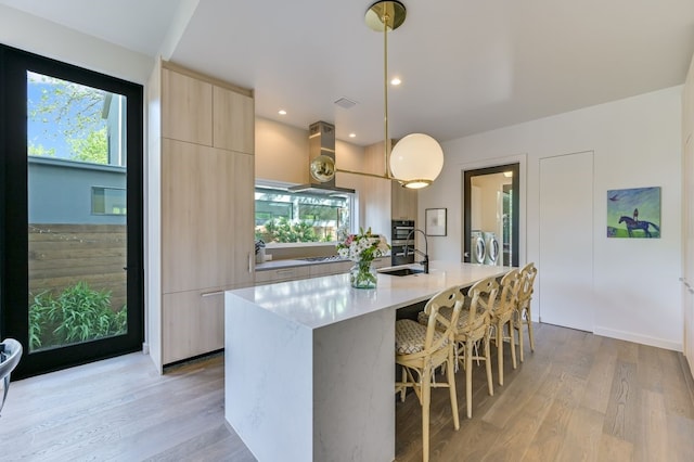 kitchen featuring washer and clothes dryer, a center island with sink, light brown cabinets, a wealth of natural light, and pendant lighting