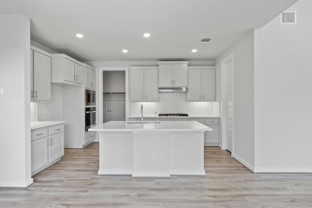 kitchen with appliances with stainless steel finishes, an island with sink, sink, white cabinets, and light hardwood / wood-style flooring