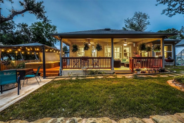 back house at dusk featuring ceiling fan, a patio area, a yard, and a hot tub