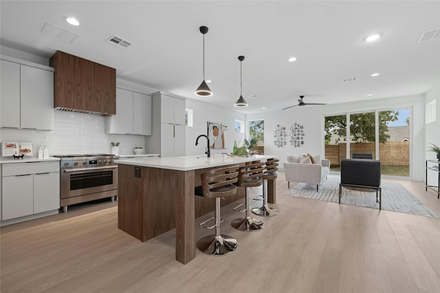 kitchen featuring stainless steel range, white cabinetry, hanging light fixtures, a kitchen island with sink, and light wood-type flooring