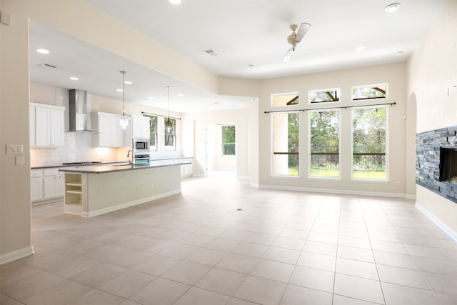 kitchen with a stone fireplace, hanging light fixtures, wall chimney range hood, an island with sink, and white cabinetry