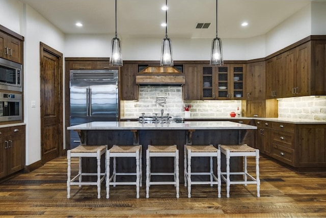 kitchen featuring backsplash, a kitchen island with sink, a kitchen breakfast bar, built in appliances, and dark brown cabinets