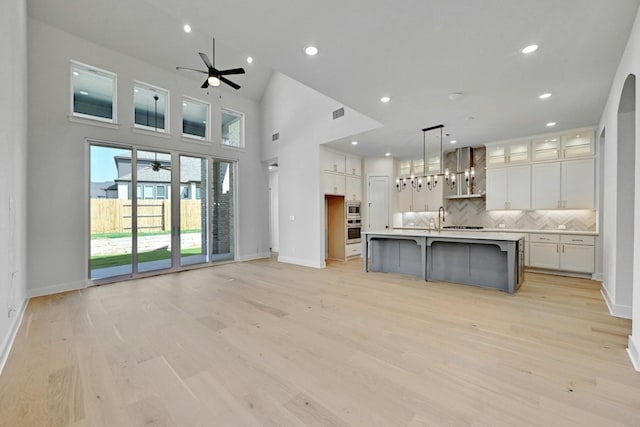 kitchen featuring wall chimney range hood, light hardwood / wood-style flooring, hanging light fixtures, a center island with sink, and white cabinetry