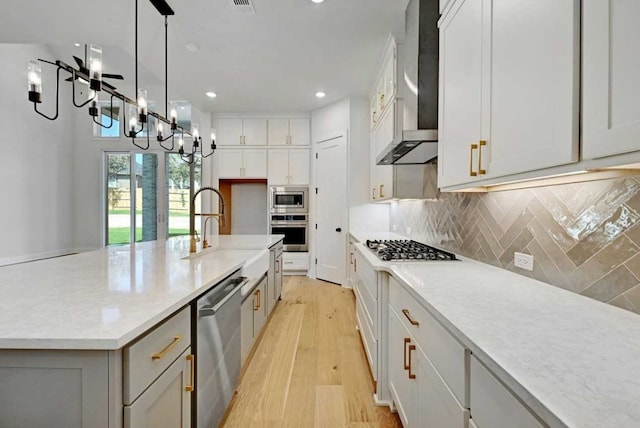 kitchen with wall chimney exhaust hood, an island with sink, hanging light fixtures, appliances with stainless steel finishes, and light hardwood / wood-style floors