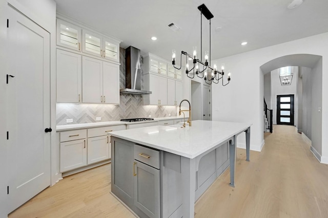 kitchen featuring hanging light fixtures, light hardwood / wood-style floors, wall chimney exhaust hood, white cabinets, and a kitchen island with sink