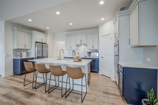 kitchen featuring an island with sink, sink, blue cabinetry, stainless steel refrigerator, and light wood-type flooring