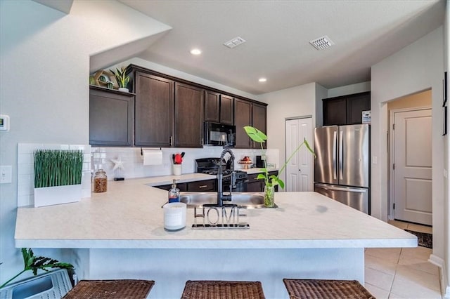 kitchen featuring dark brown cabinets, kitchen peninsula, a kitchen bar, backsplash, and stainless steel refrigerator