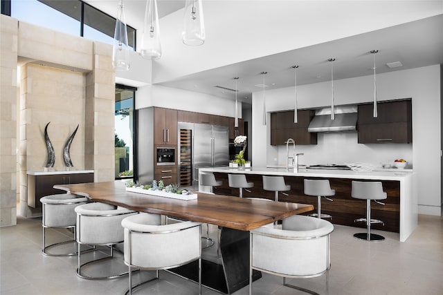 kitchen featuring appliances with stainless steel finishes, a towering ceiling, wall chimney exhaust hood, dark brown cabinets, and hanging light fixtures