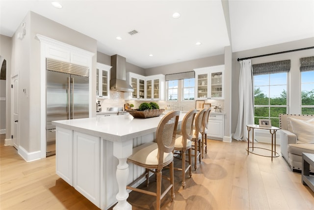 kitchen featuring wall chimney exhaust hood, a kitchen island, light hardwood / wood-style flooring, stainless steel built in fridge, and white cabinets