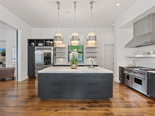 kitchen with range with two ovens, backsplash, wall chimney exhaust hood, a kitchen island with sink, and dark hardwood / wood-style flooring