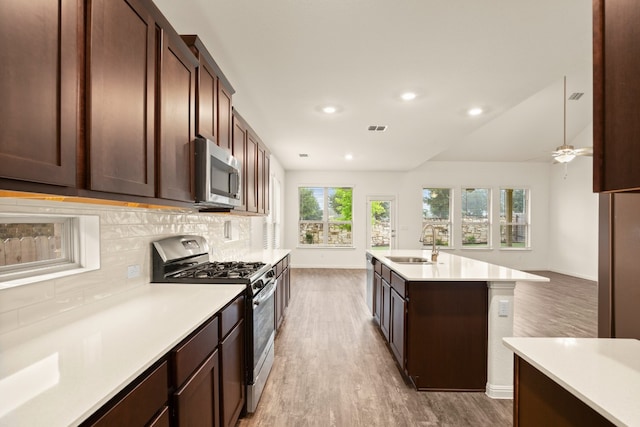 kitchen with sink, a center island with sink, decorative backsplash, appliances with stainless steel finishes, and wood-type flooring