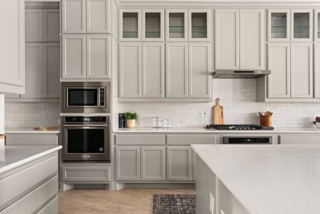 kitchen with light stone counters, under cabinet range hood, appliances with stainless steel finishes, tasteful backsplash, and glass insert cabinets