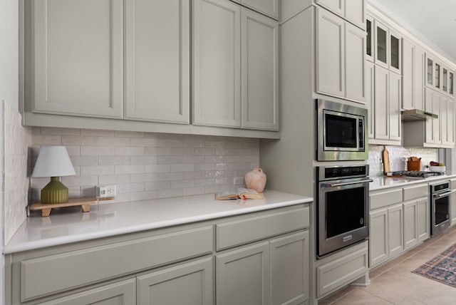 kitchen featuring light tile patterned floors, light countertops, appliances with stainless steel finishes, decorative backsplash, and glass insert cabinets