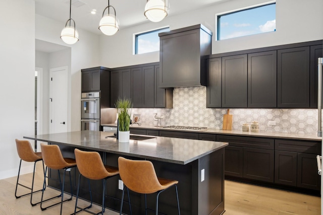 kitchen featuring appliances with stainless steel finishes, a kitchen island with sink, decorative backsplash, wall chimney range hood, and decorative light fixtures