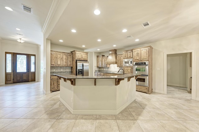 kitchen with a breakfast bar area, stainless steel appliances, a center island with sink, and custom range hood